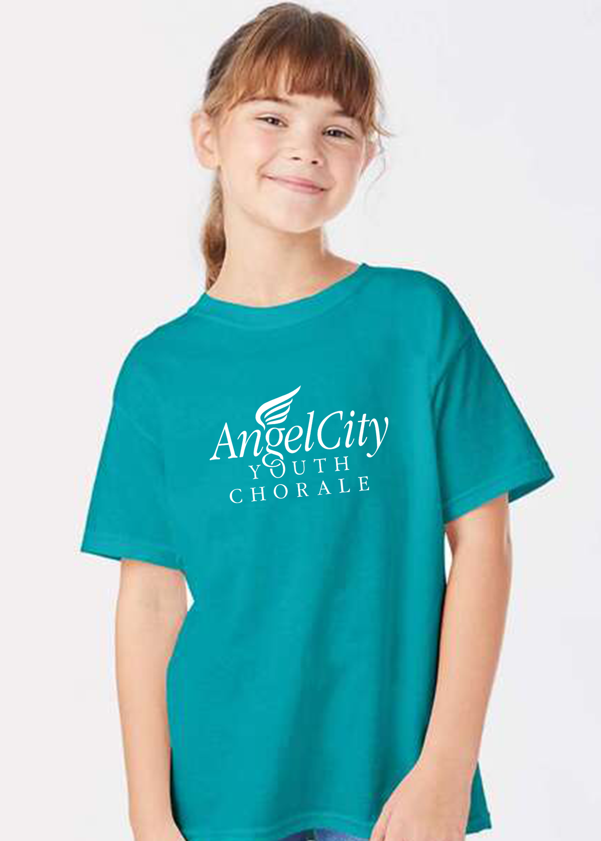 A young girl with brown hair in a ponytail smiling while wearing a teal short-sleeve t-shirt with 'Angel City Youth Chorale' printed in white on the front, set against a plain light gray background.